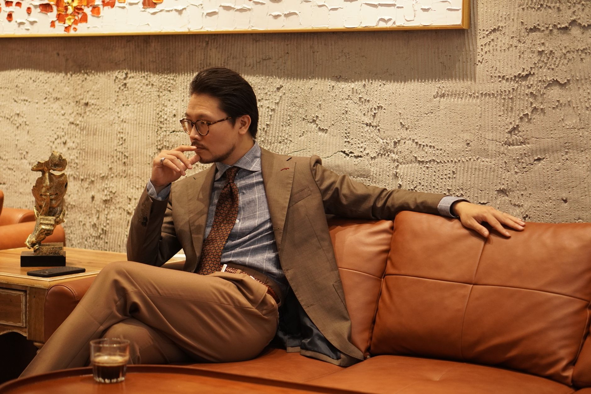 Well-dressed man in a suit and glasses sitting on a leather couch, appearing thoughtful.