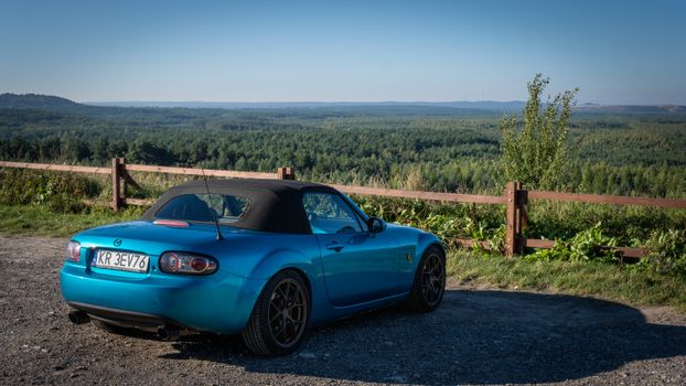 A blue Mazda MX-5 sports car parked on a scenic hill with a vast forest view.