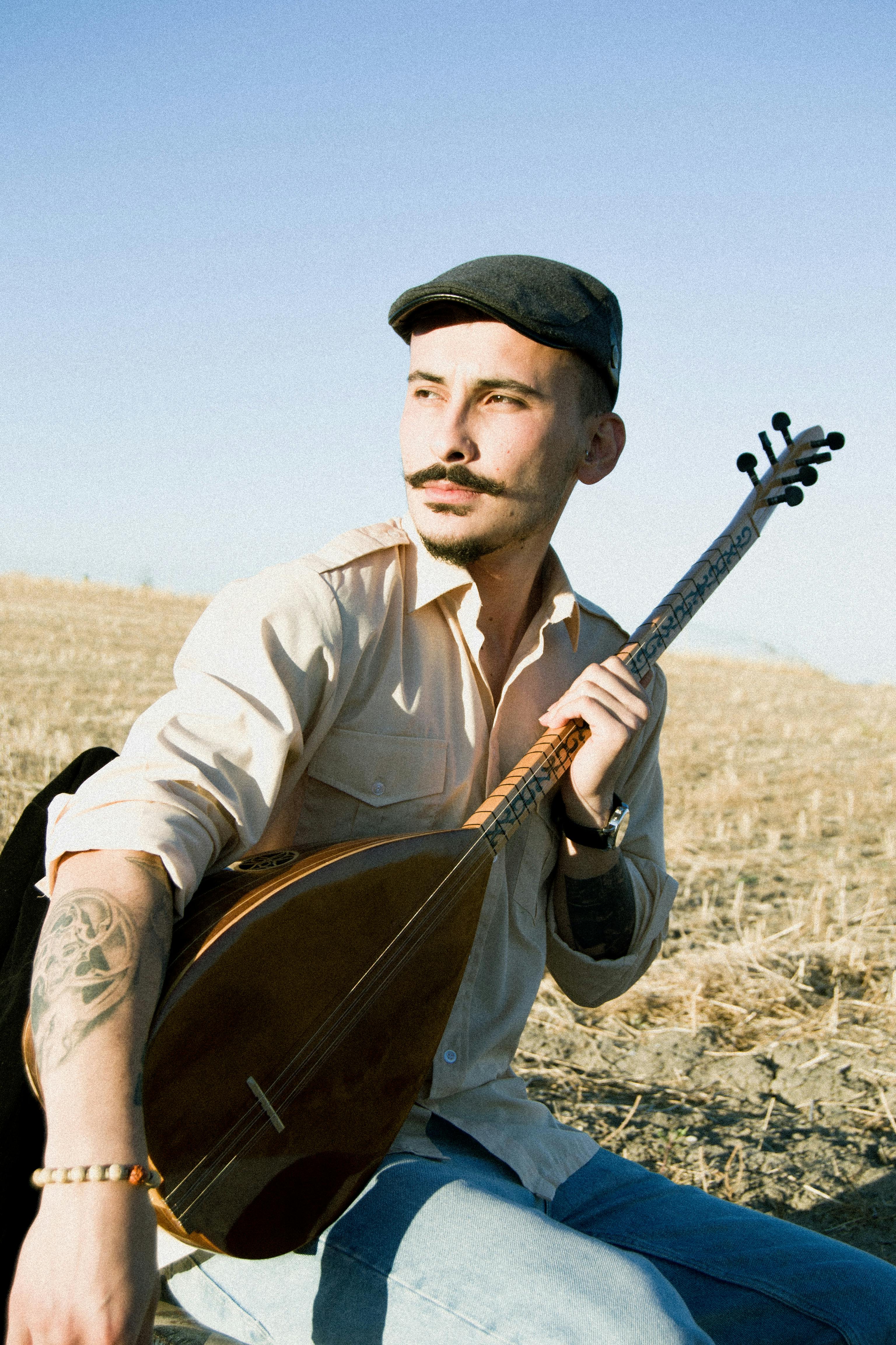 Musician with tattoos playing traditional string instrument in a rural field.