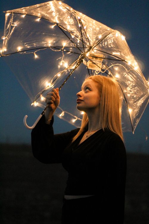 Free A young woman gazes upward, holding a glowing umbrella against a dark night sky. Stock Photo