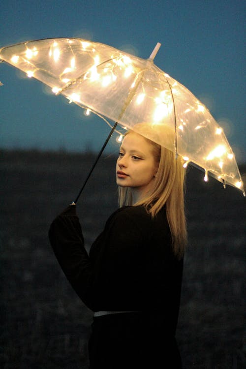 Young Blonde Woman in Black Blouse Posing under Umbrella Decorated with LEDs