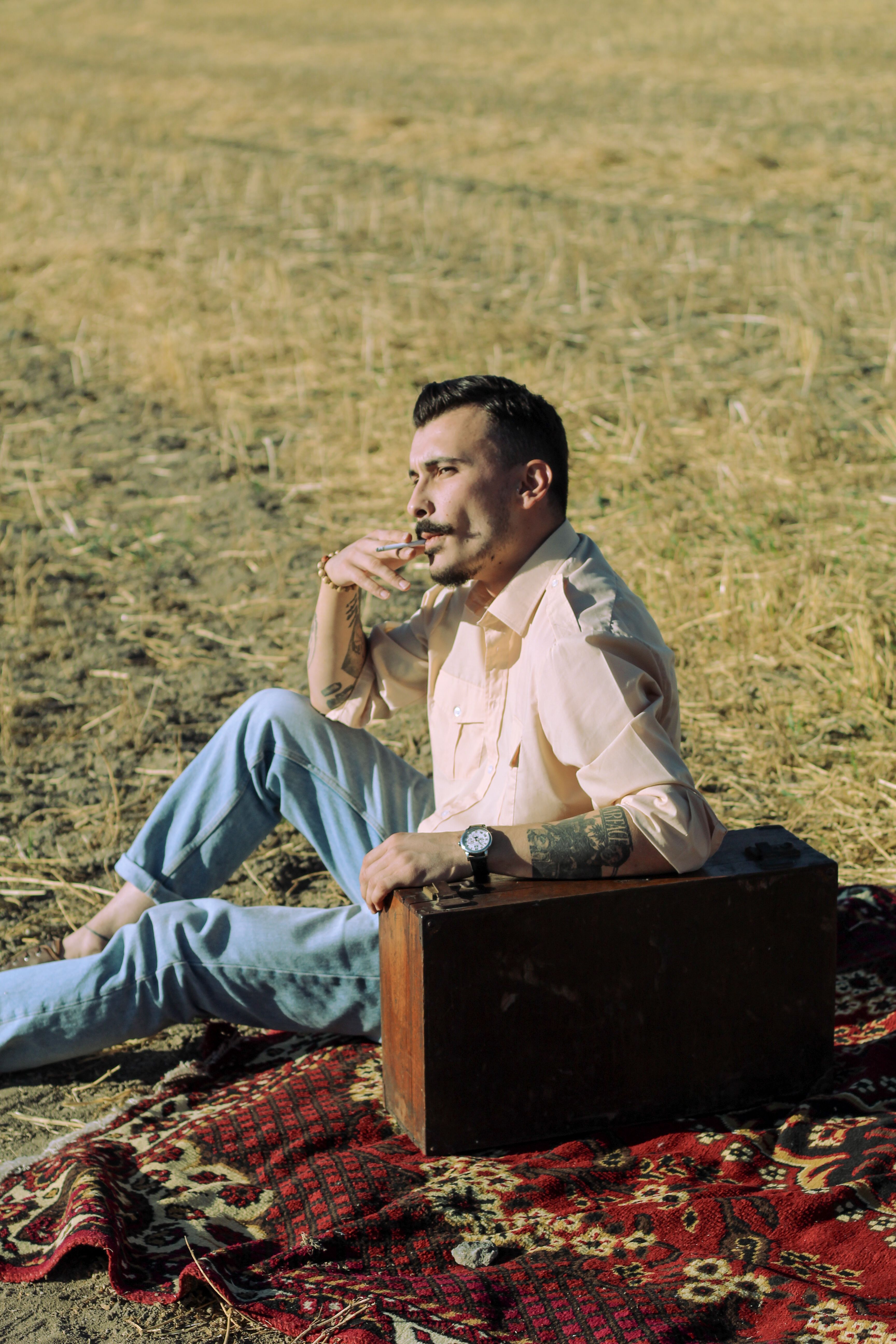 A man with a mustache sits on a rug in a rural field, smoking and holding a suitcase.