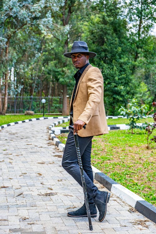 Model in a Beige Blazer and a Hat Standing with a Cane on a Walkway in the Park