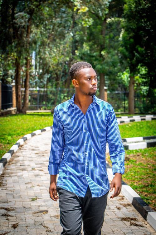 Free A young man in a striped shirt walks on a sunlit pathway through a green park. Stock Photo