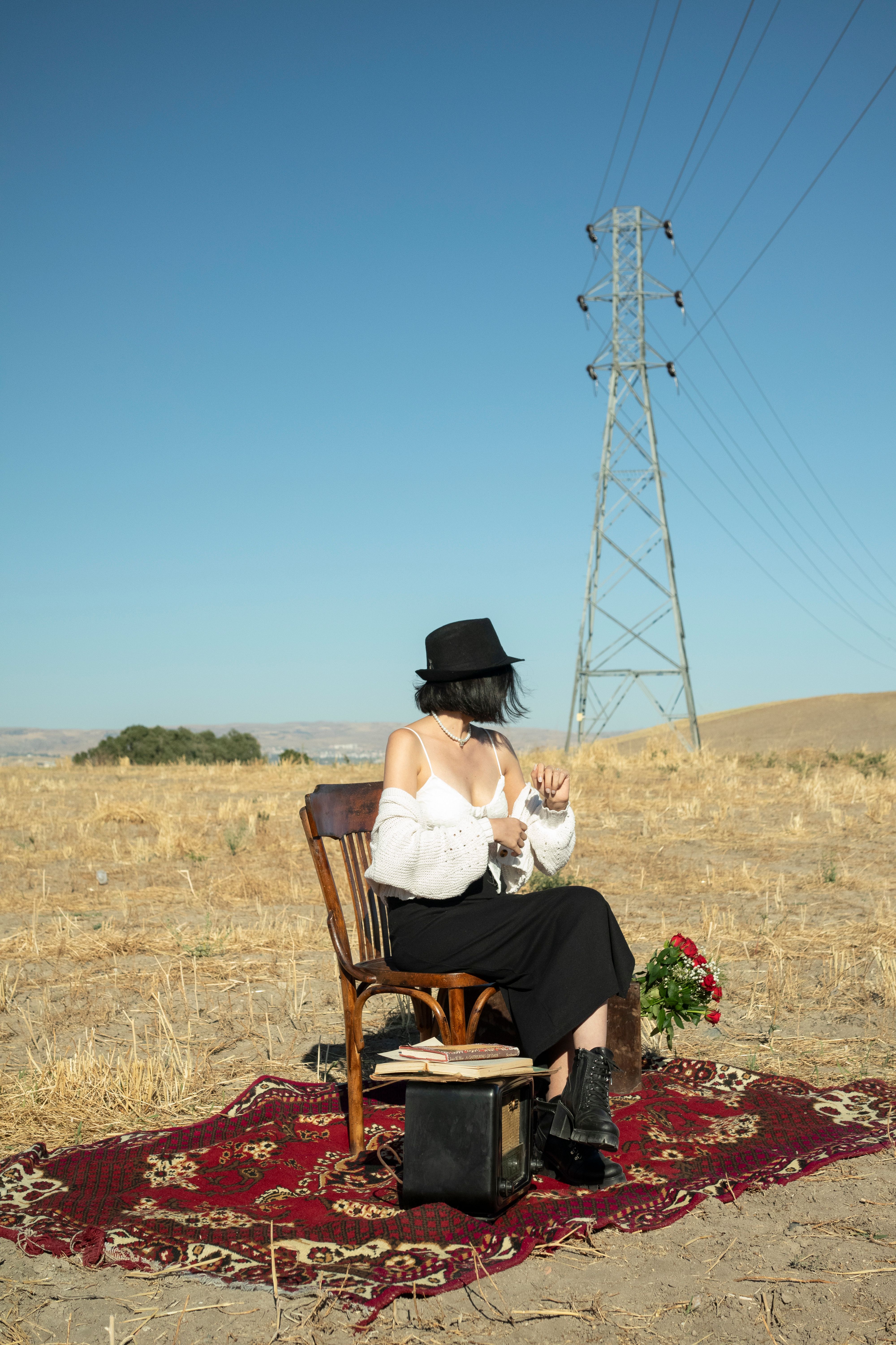 Stylish woman in hat poses outdoors on a rug, embracing a retro vibe in rural scenery.