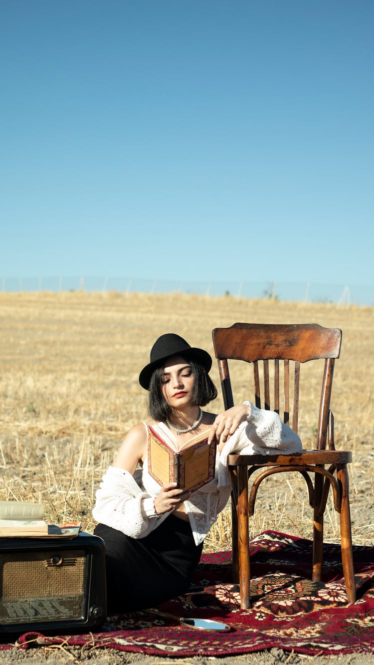 Woman Reading A Book Sitting On The Carpet Among The Fields With Her Hand Resting On The Chair