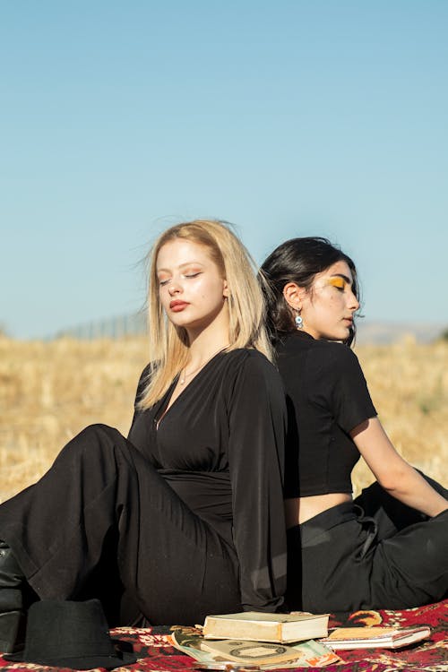 Two Young Women in Black Outfits Sitting on a Field in Sunlight 