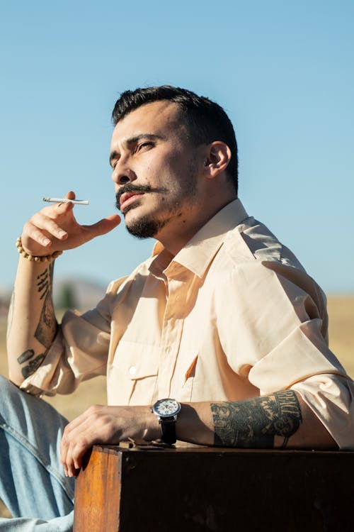 Man in a Beige Shirt Sitting in a Field Smoking Cigarette