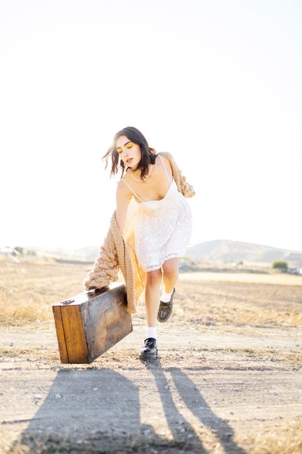 A young woman in a retro dress poses with a vintage suitcase in a sunny outdoor setting.
