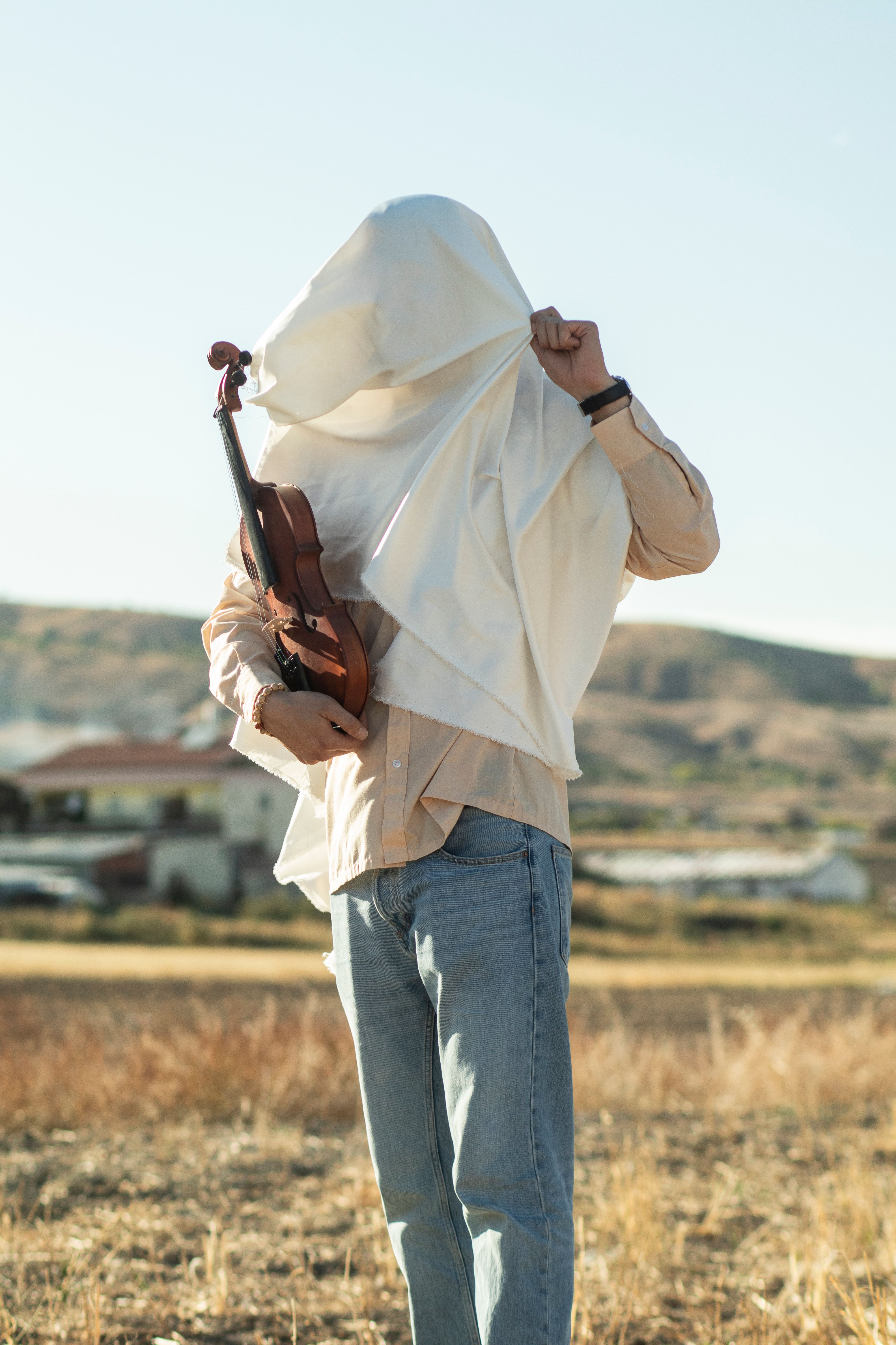 A person partially covered by a cloth holds a violin outdoors in a sunny field.