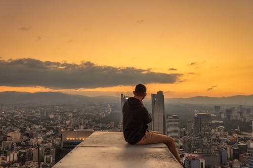 Free Man Sitting on a Rooftop and Contemplating the Cityscape with a Yellow Sky Stock Photo