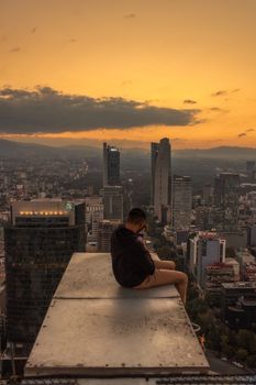 A man sitting on a rooftop edge at sunset, overlooking a sprawling city skyline.
