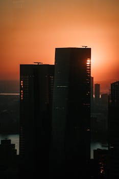 Silhouetted skyscrapers against an orange sunset sky create a dramatic urban scene.
