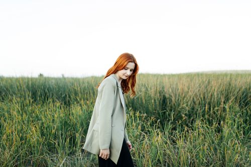 Free A young redheaded woman in a blazer walks thoughtfully through a green field. Stock Photo