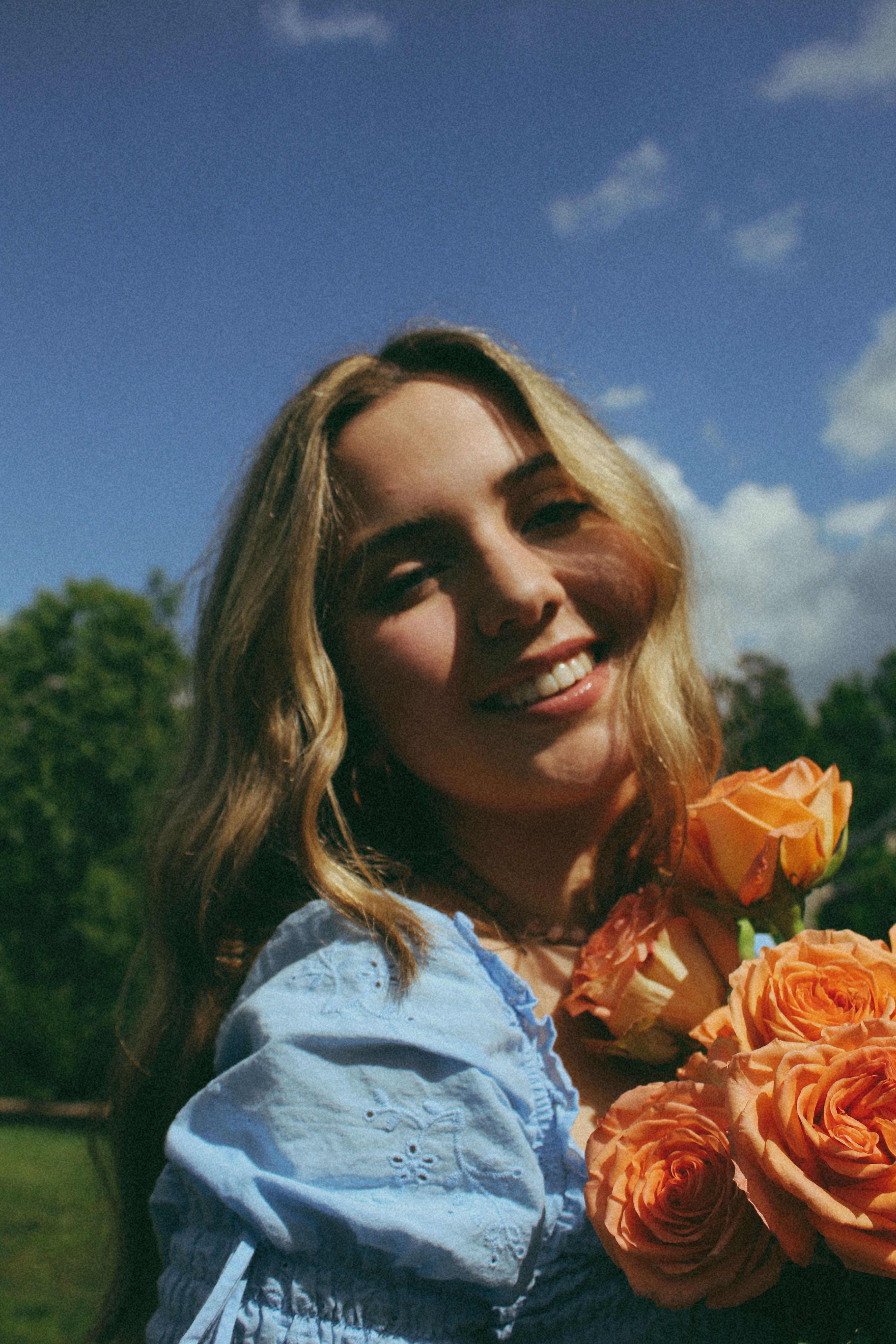 Smiling woman holding an orange rose bouquet in a sunlit park.