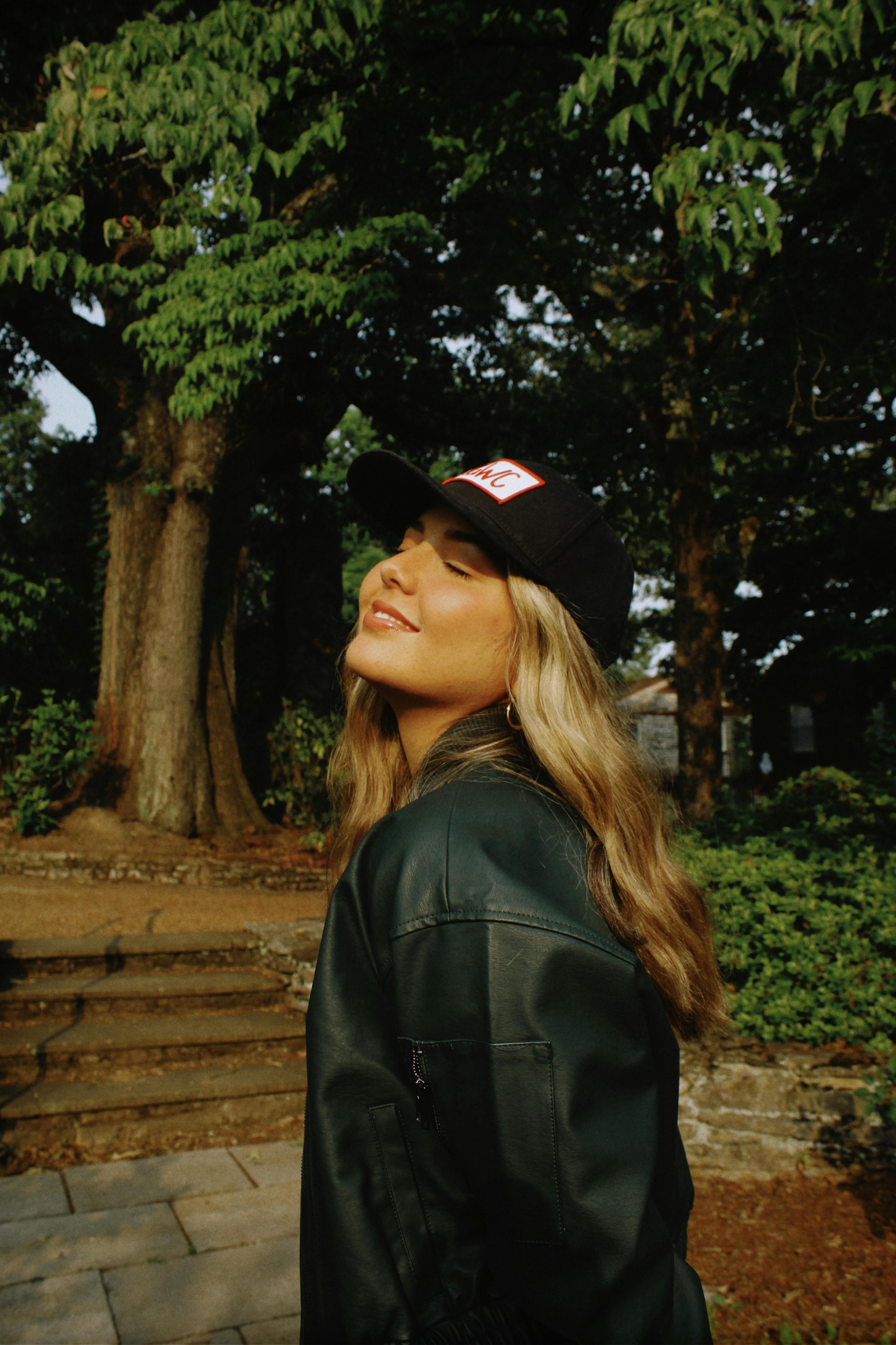 A smiling blonde woman with closed eyes enjoying the outdoors in a summer park.