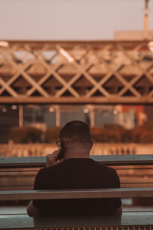 Free Rear view of a man sitting on a bench, speaking on a smartphone in a city environment. Stock Photo