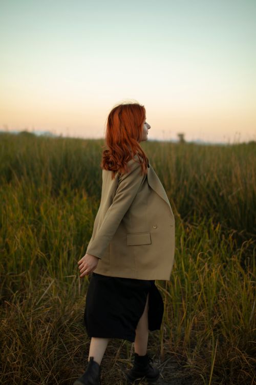 Free A red-haired woman in a blazer and skirt walks through a grass field at dawn. Stock Photo