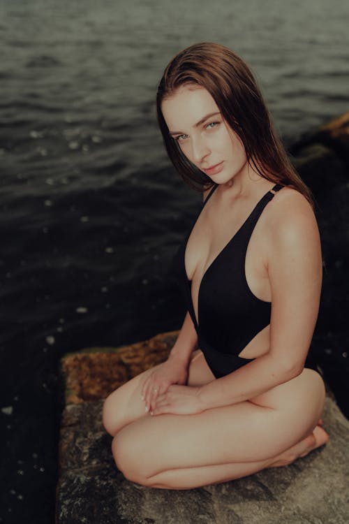A beautiful young woman in a black swimsuit sitting on a rock