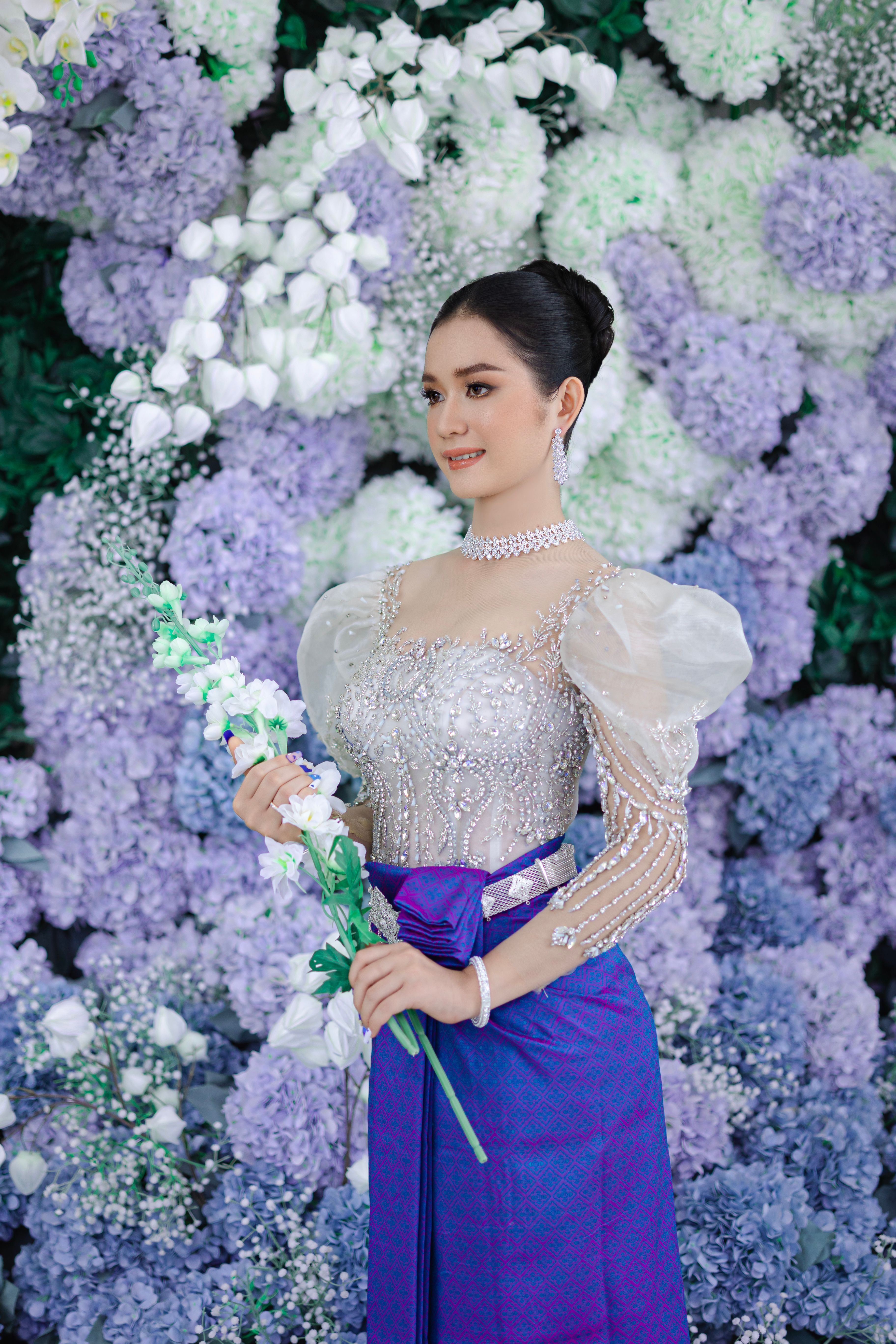 Asian woman elegantly posing in traditional attire amidst a floral backdrop.