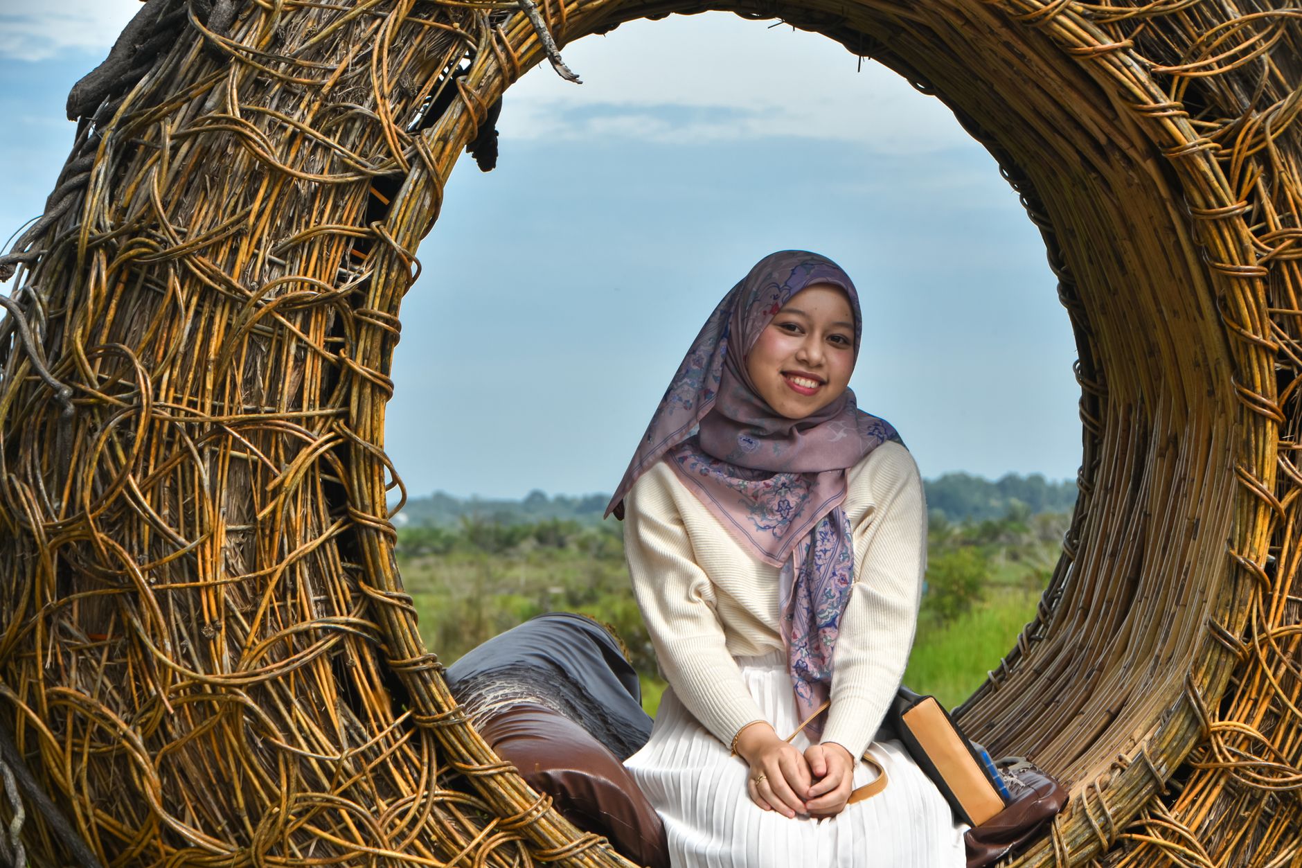 A young woman in a headscarf smiles while sitting in a wicker circle in a rural setting.