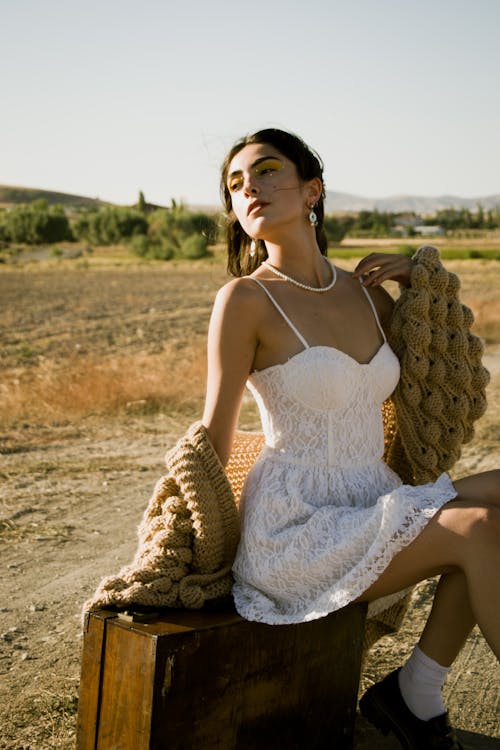 Model in a White Lace Dress and Cardigan Sitting on an Old Wooden Suitcase