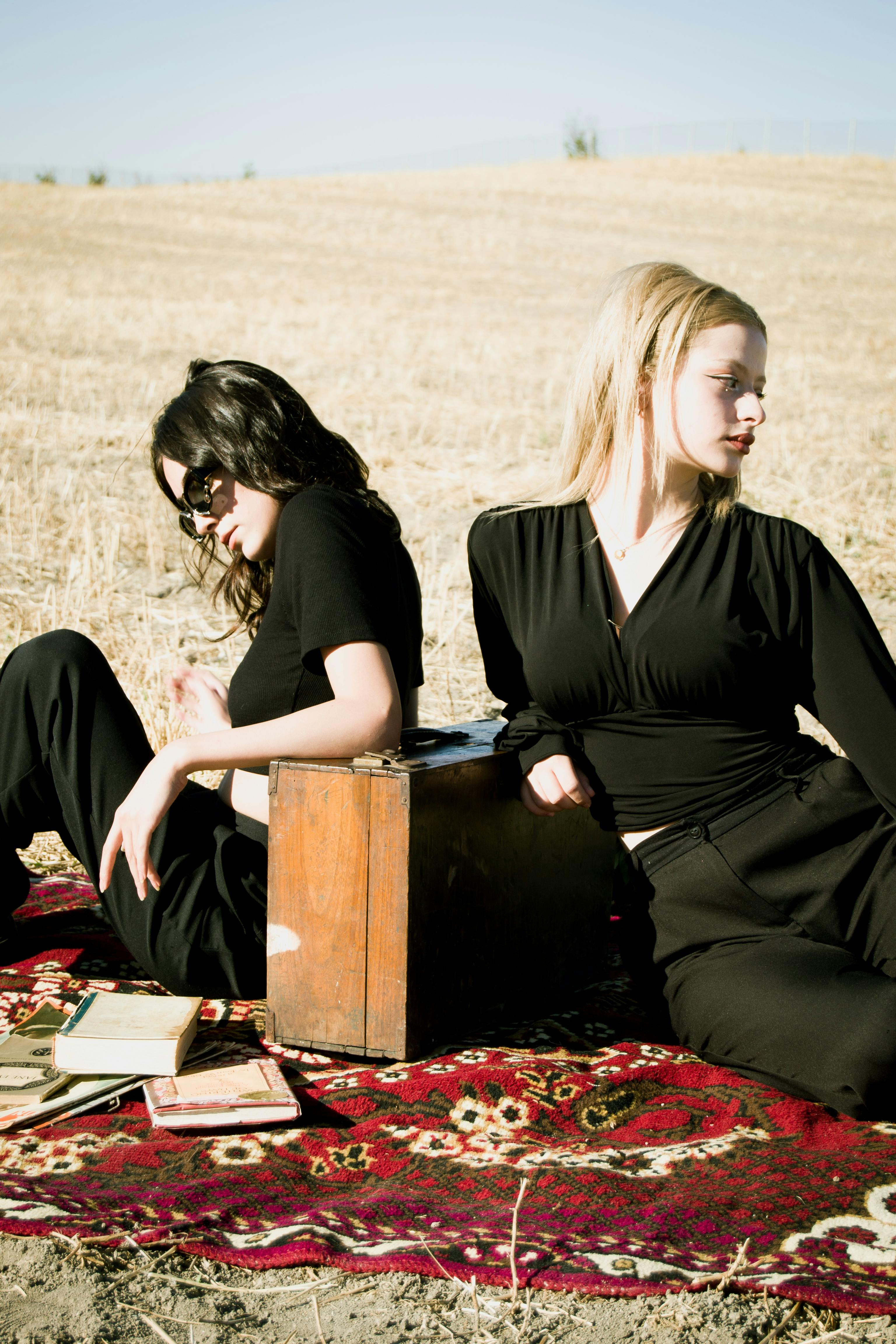 Models Dressed in Black Sitting Next to an Old Suitcase on a Carpet Spread  in a Field · Free Stock Photo