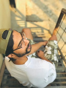 A young man in casual attire, relaxing outdoors on stairs, wearing a cap and eyeglasses.