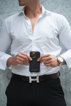 A stylish man in a white shirt holding a smartphone against a concrete wall backdrop.