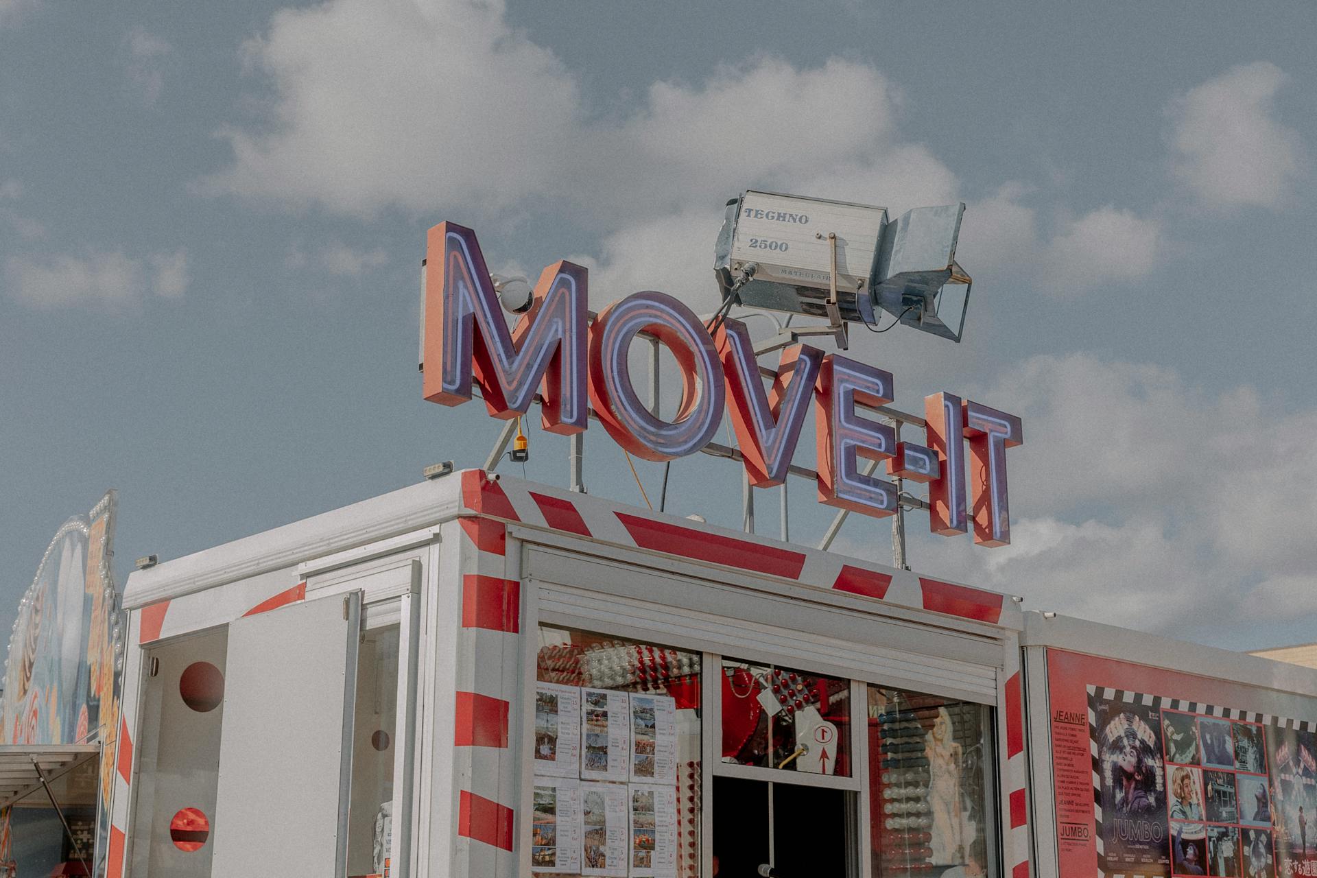 Vibrant ticket booth with neon 'MOVE-IT' sign at a sunny carnival setting.