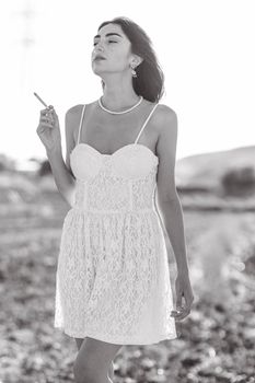 Black and white photo of a woman in a white lace dress smoking outdoors.