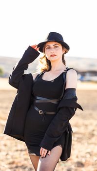 Portrait of a fashionable woman in black attire and hat standing outdoors in rural setting.