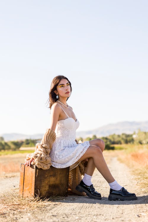 Woman in White Dress Sitting on Suitcase on Dirt Road