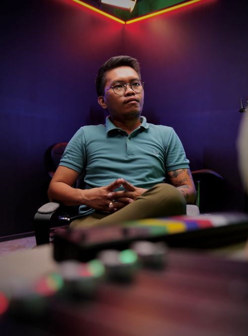 Free Portrait of a man sitting in a neon-lit studio setting, wearing glasses and a polo shirt. Stock Photo