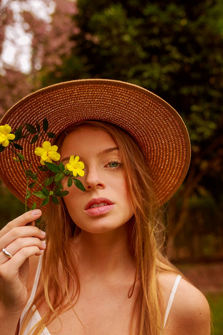 Woman In Wearing Straw Hat Holding Yellow Flowers Near Her Face