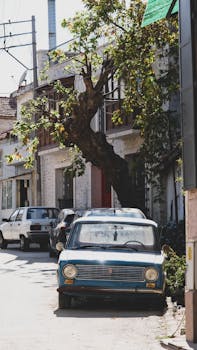 A classic car parked on a sunlit narrow street lined with trees and buildings.