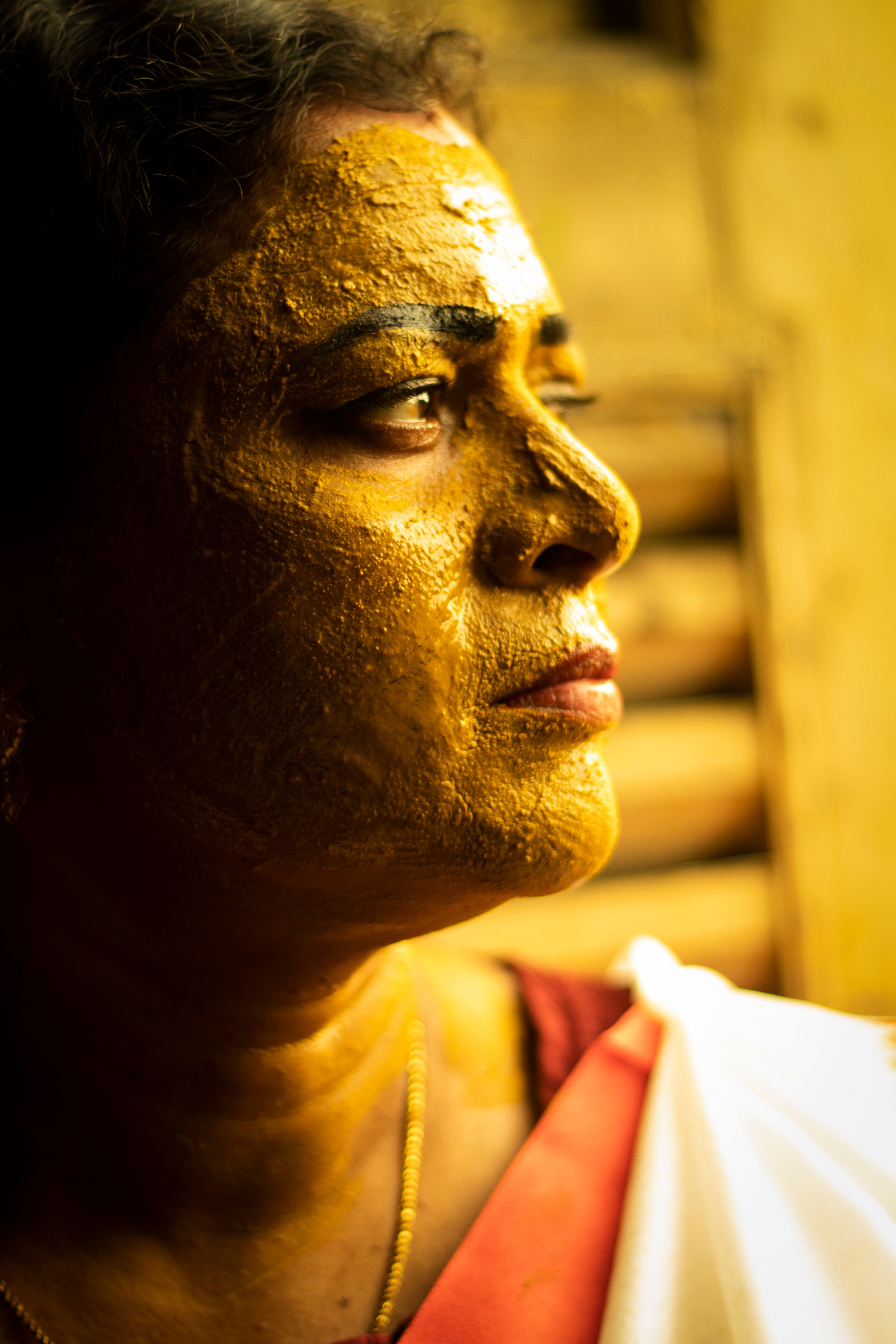 A South Asian woman with golden paint on her face gazes thoughtfully indoors, captured in a warm portrait.