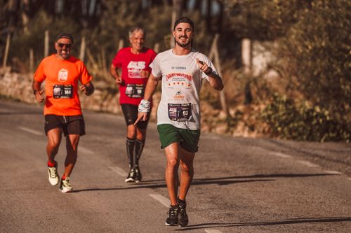 Free Three men running in a sunny marathon, wearing sportswear and smiling on an empty road. Stock Photo
