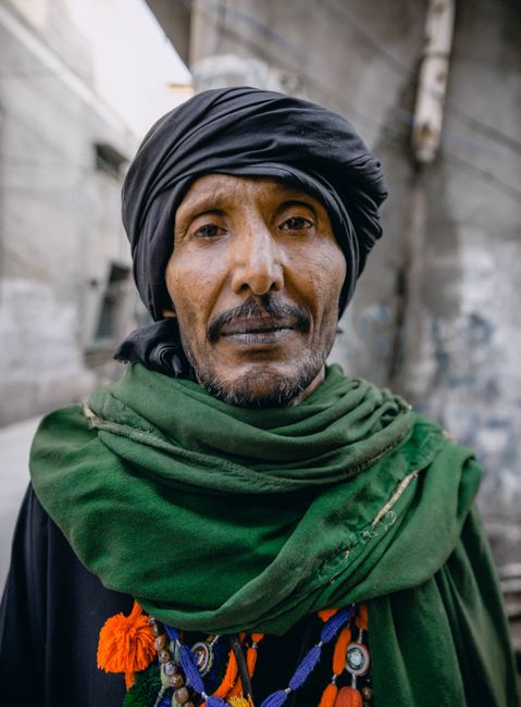 Close-up portrait of an adult man in traditional attire and green scarf outdoors.