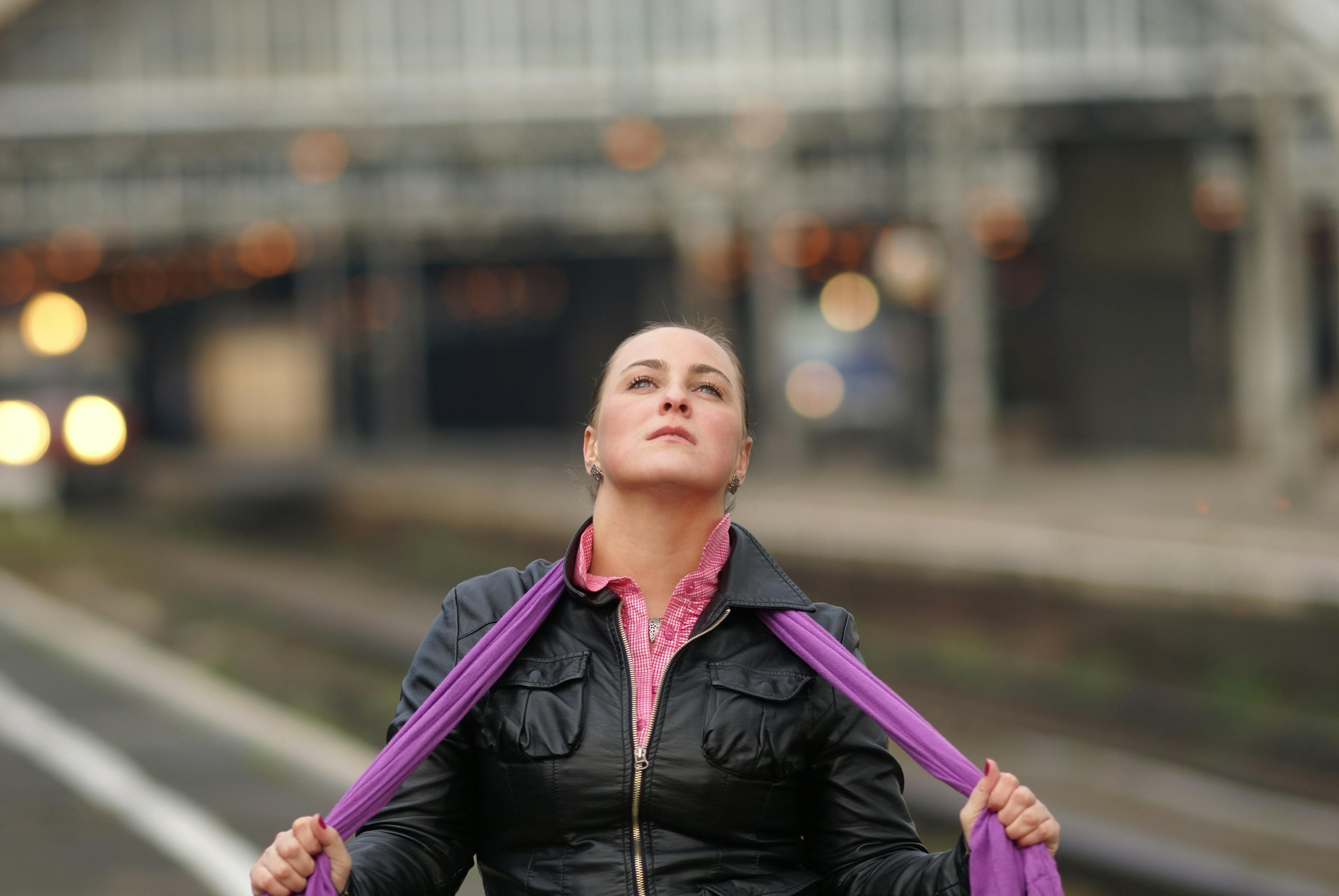 A woman wearing a leather jacket stands at a train station with a contemplative gaze.