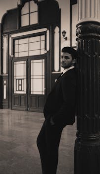 A stylish man in a black suit poses against a column inside a classic building.