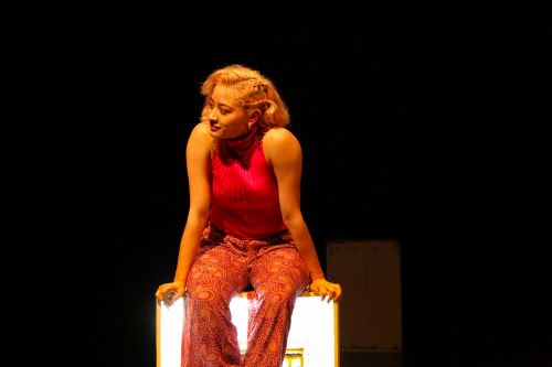 Free Blonde woman in vibrant red and patterned pants sitting confidently on a stage against a black background. Stock Photo