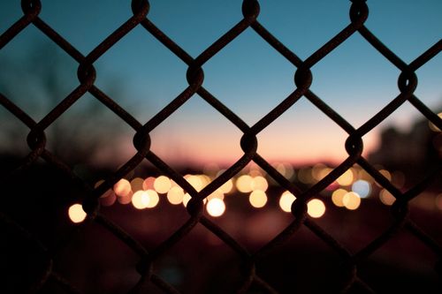 Free Chain link fence silhouette with bokeh city lights at sunset, creating a moody urban atmosphere. Stock Photo