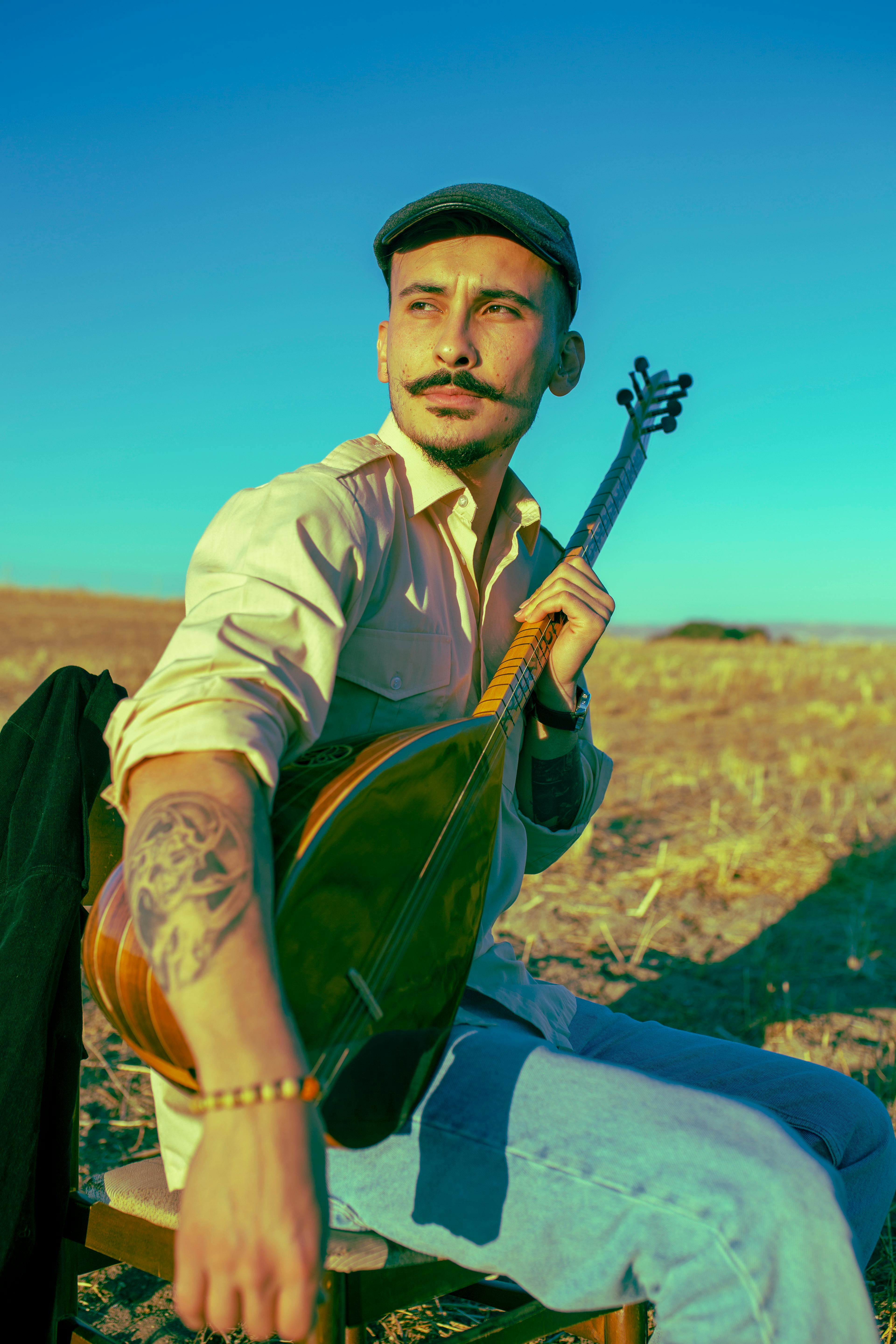 Man with mustache and cap playing a baglama in a rural field.