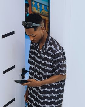 A fashionable man stands indoors using a smartphone, wearing sunglasses.
