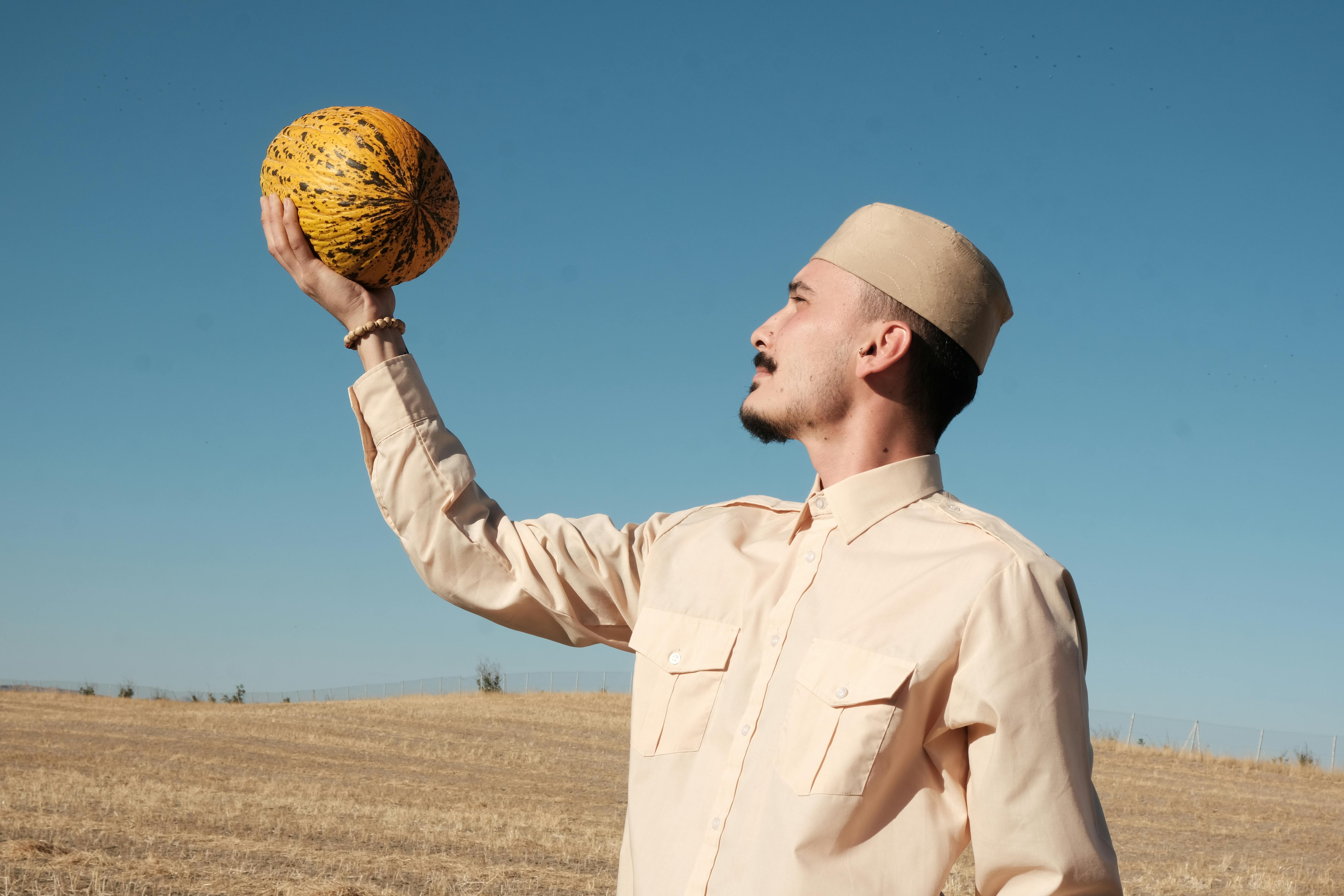 A man in a rural landscape holds a melon while looking up against a clear blue sky.