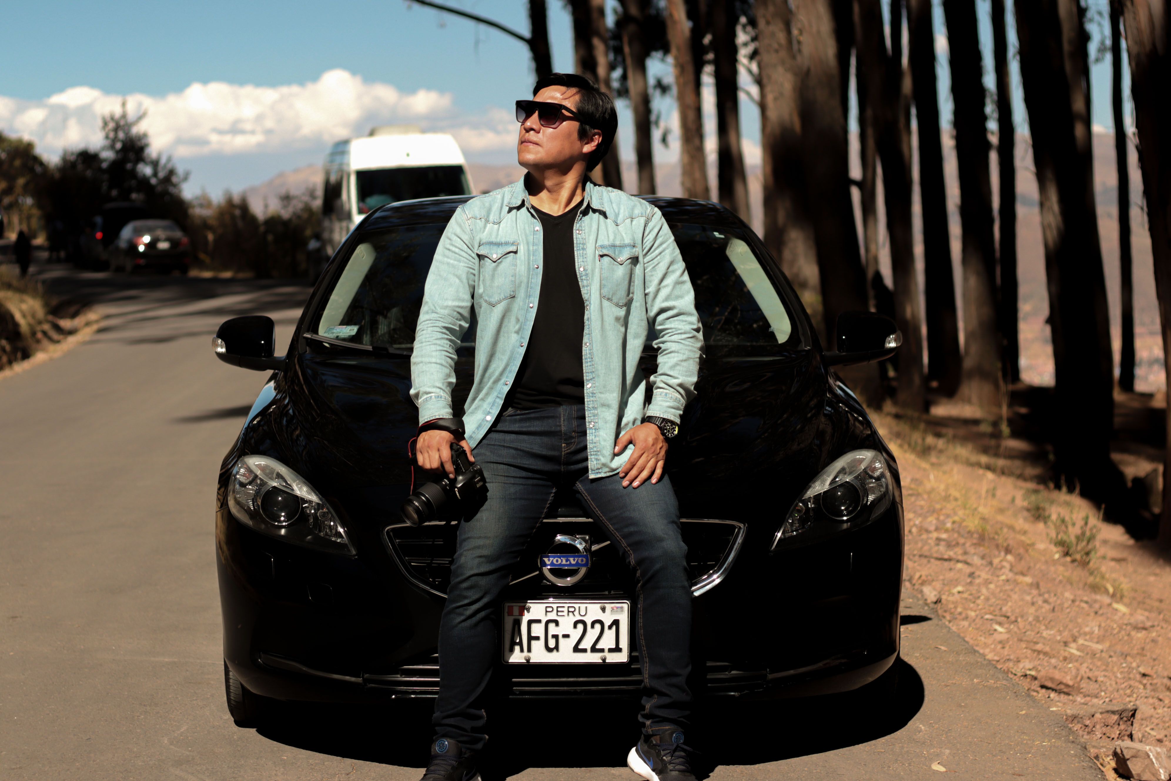 Stylish man in sunglasses leaning on a Volvo, captured on a sunlit road in Cusco, Peru. Fashion and travel vibes.