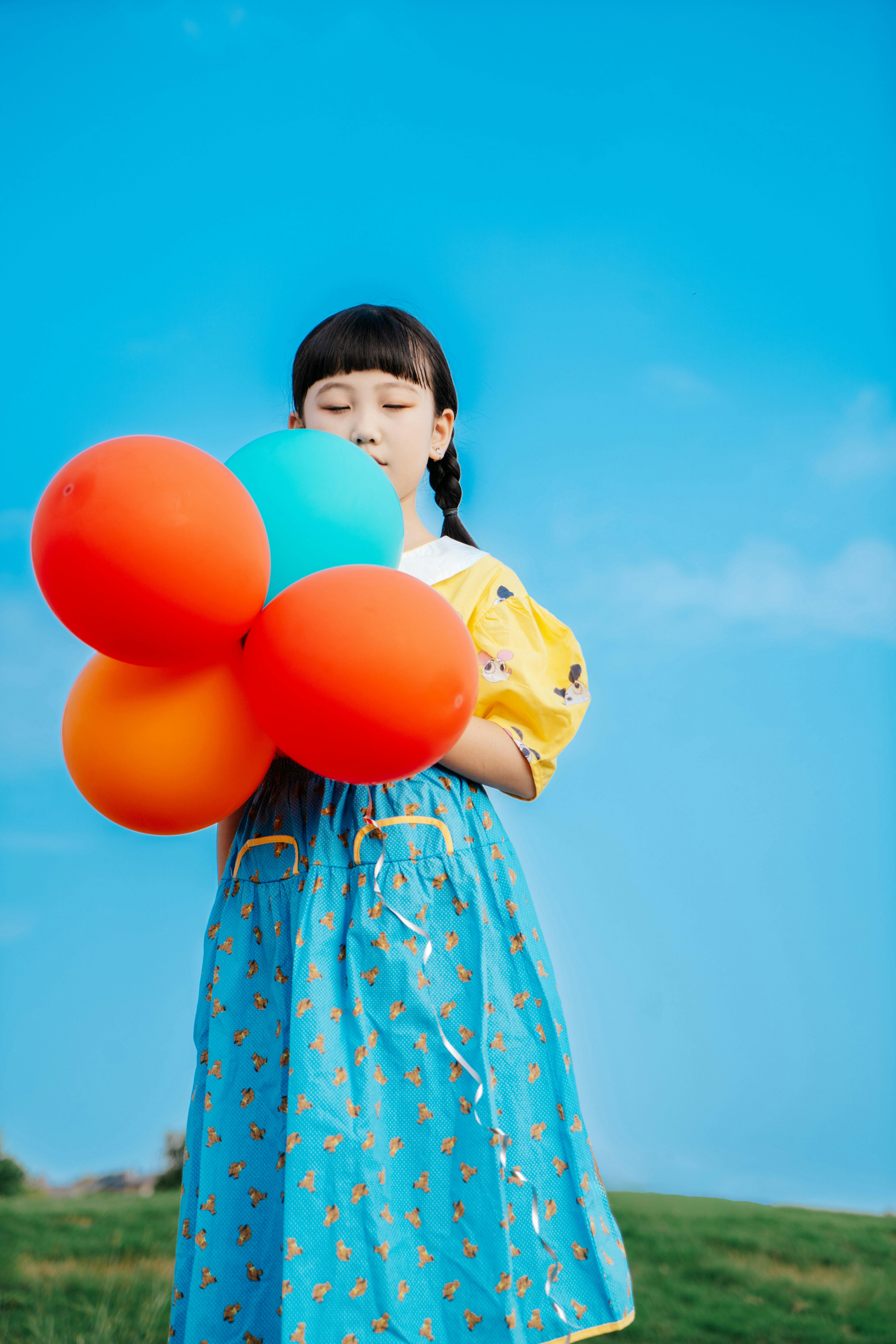 A young girl in a vibrant dress holds colorful balloons against a clear blue sky.