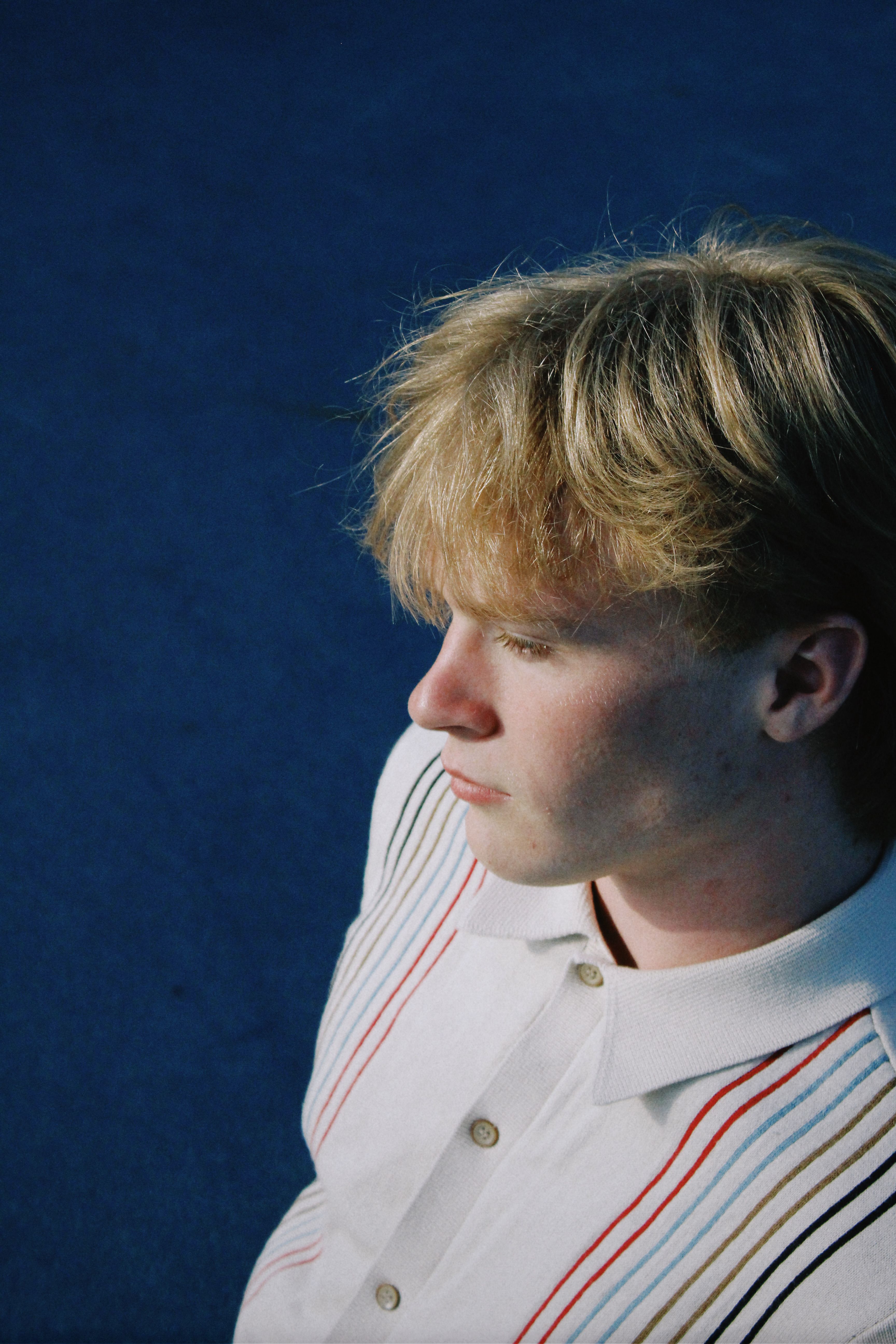 Portrait of a young man with blonde hair wearing a striped shirt against a blue backdrop.