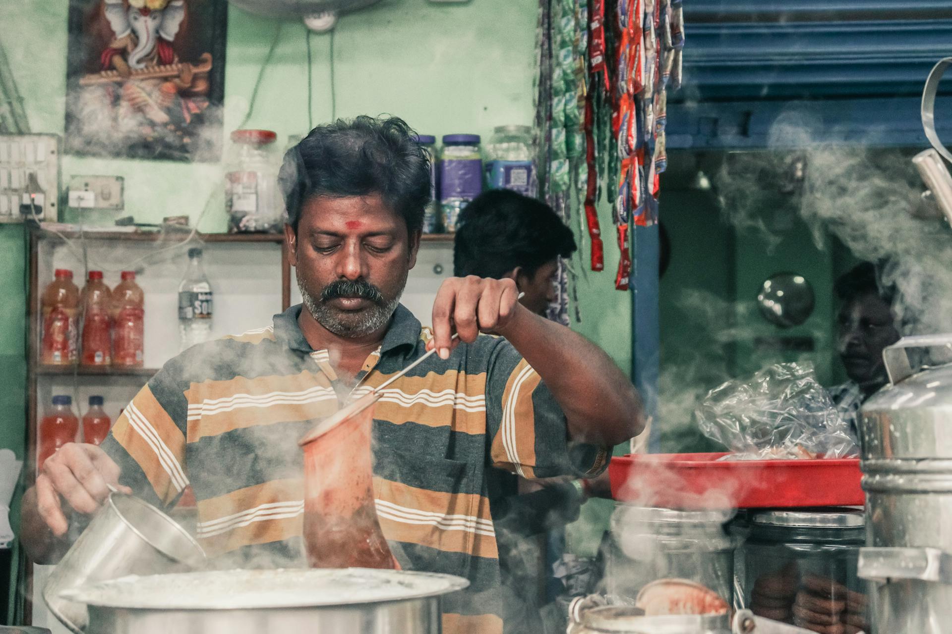 A man making traditional masala chai in an Indian tea stall, surrounded by steam and kitchen utensils.